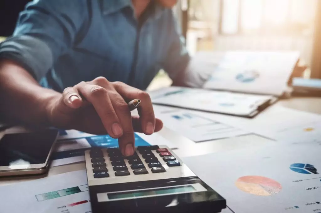 Person sitting at desk putting numbers into a calculator surrounded by sheets of financial paper with pie charts on them